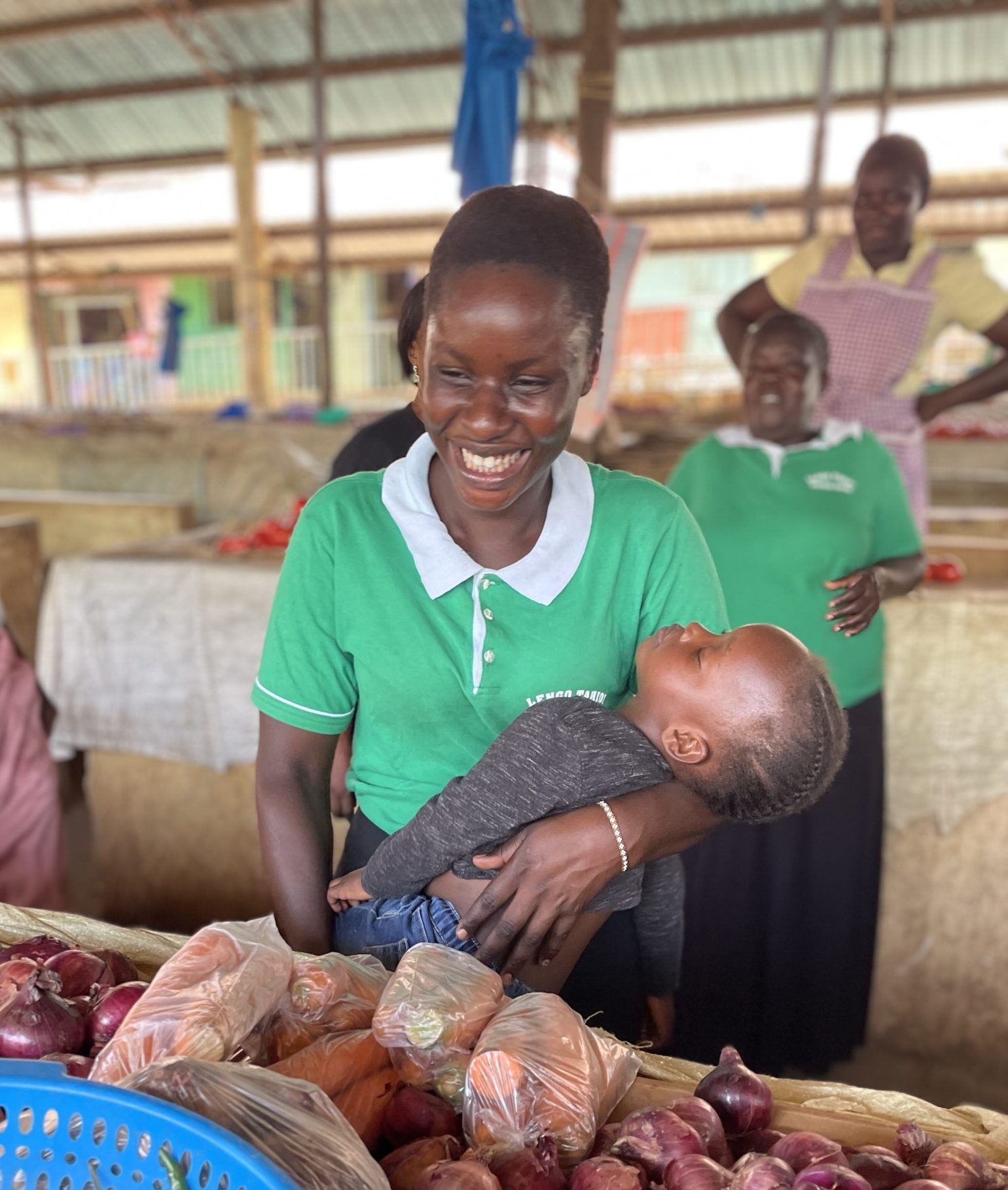 Lavinda with daughter in her arms, in front of vegetable stall