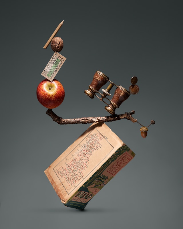 A collection of items balance on top of a map, the items include an apple, binoculars, a pencil and more
